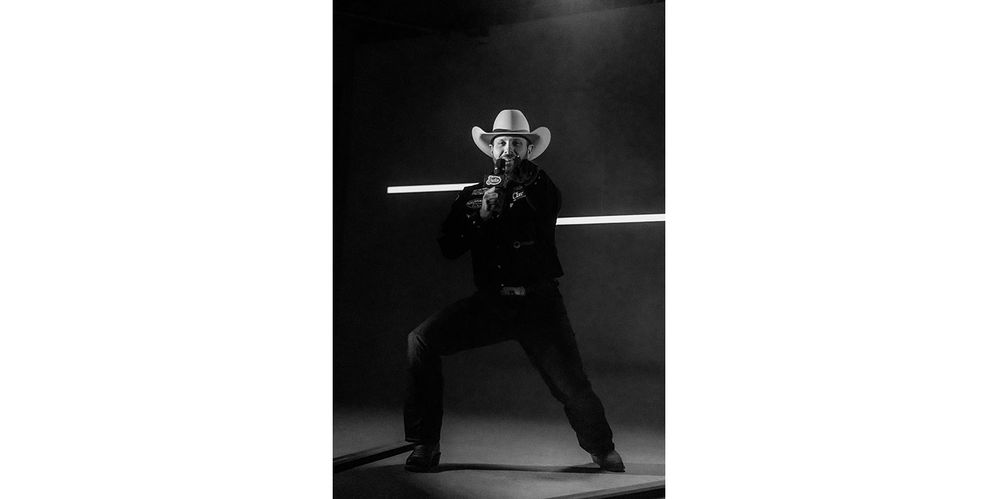 Anthony Lucia vistiendo una camisa negra y un sombrero de vaquero, sosteniendo un micrófono y posando mientras apunta a la cámara.